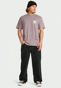 Απαλή μωβ βαμβακερή μπλούζα με σχέδιο στο στήθος, σε συνδυασμό με μαύρο φαρδύ παντελόνι και μαύρες-άσπρες αθλητικές παπούτσια, φορεμένα με ένα μαύρο σκουφάκι.