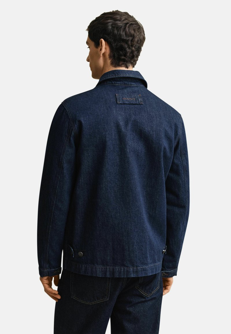 Uomo con giacca di jeans blu scuro e jeans abbinati, visto di spalle su uno sfondo bianco uniforme.