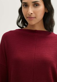 Bordeauxfarbener, strukturierter Pullover mit Bootsausschnitt, der ein geripptes Muster und einen lockeren Schnitt aufweist. Glattes, gestricktes Material.