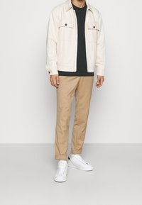 Veste beige clair avec deux poches avant, t-shirt noir en dessous, pantalon marron clair et baskets blanches avec des accents subtils.