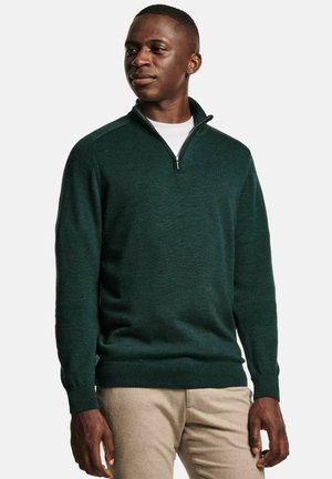 Pull en maille vert foncé avec un col zippé à quart, des poignets et un ourlet côtelés. Porté par-dessus une chemise blanche, assorti avec un pantalon beige clair.