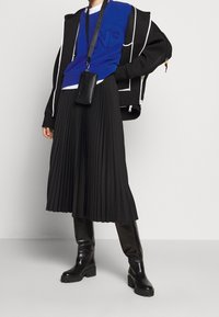 Falda negra plisada, suéter azul con logo, chaqueta negra de gran tamaño con cremallera y botas altas negras. Bolso negro cruzado con correa.