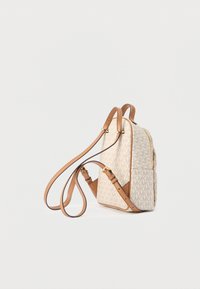 Petit sac à dos beige avec des bretelles réglables marron clair et des poches latérales, présentant un motif monogramme MK et une fermeture éclair dorée.