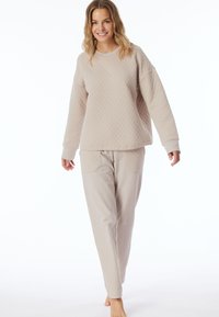 Beige gewatteerde pullover met lange mouwen, gecombineerd met bijpassende lichtgewicht broek met zakken en een ontspannen pasvorm, tegen een witte achtergrond.