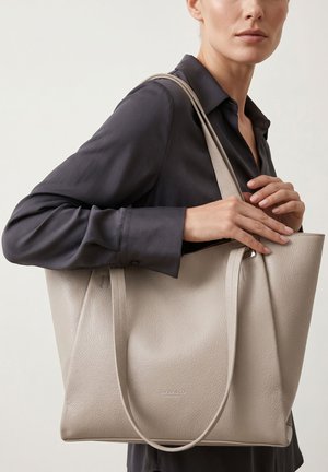 Vrouw in donker overhemd houdt een grote gestructureerde beige leren tas met dubbele schouderbanden tegen een effen achtergrond.