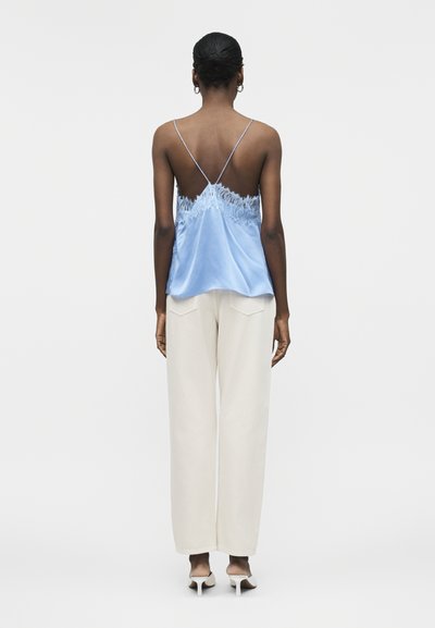 Γυναίκα όρθια, φοράει ανοιχτό μπλε καλοκαιρινή μπλούζα με δαντέλα και λεπτές τιράντες και εκρού παντελόνα με φαρδιά πόδια, εμφανιζόμενη από πίσω.