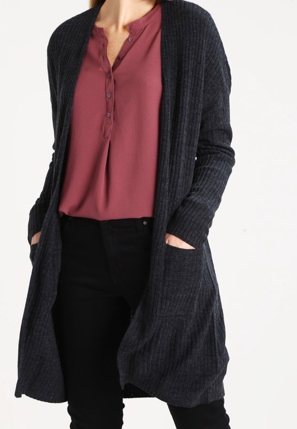 Donna con un lungo cardigan a coste scuro sopra una blusa mauve con bottoni e pantaloni neri, mani nelle tasche del cardigan.