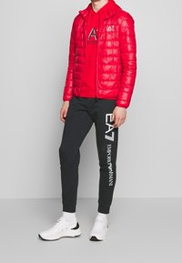 Czerwona kurtka puchowa o błyszczącej fakturze, noszona na czerwonym hoodie i czarnych dresach. Dresy mają białe logo nadrukowane na boku. Białe sneakersy.