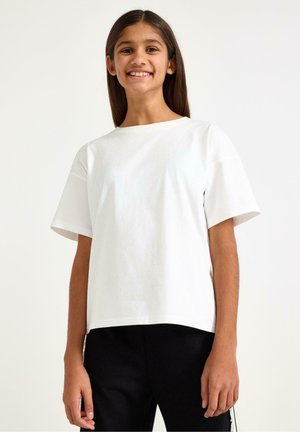 Smilende ung pige med langt mørkt hår iført en hvid T-shirt med korte ærmer og sorte bukser mod en ensfarvet lyse baggrund.