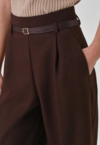 Frauenhand in der Tasche von hoch taillierter, dunkelbrauner, plissierter Hose mit schmalem dunkelbraunem Gürtel und beigem, geripptem Oberteil, das eingesteckt ist.