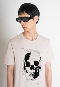 Camiseta beige con un gráfico de calavera negra con detalles texturales. El modelo lleva gafas de sol rectangulares negras, destacando un diseño moderno.