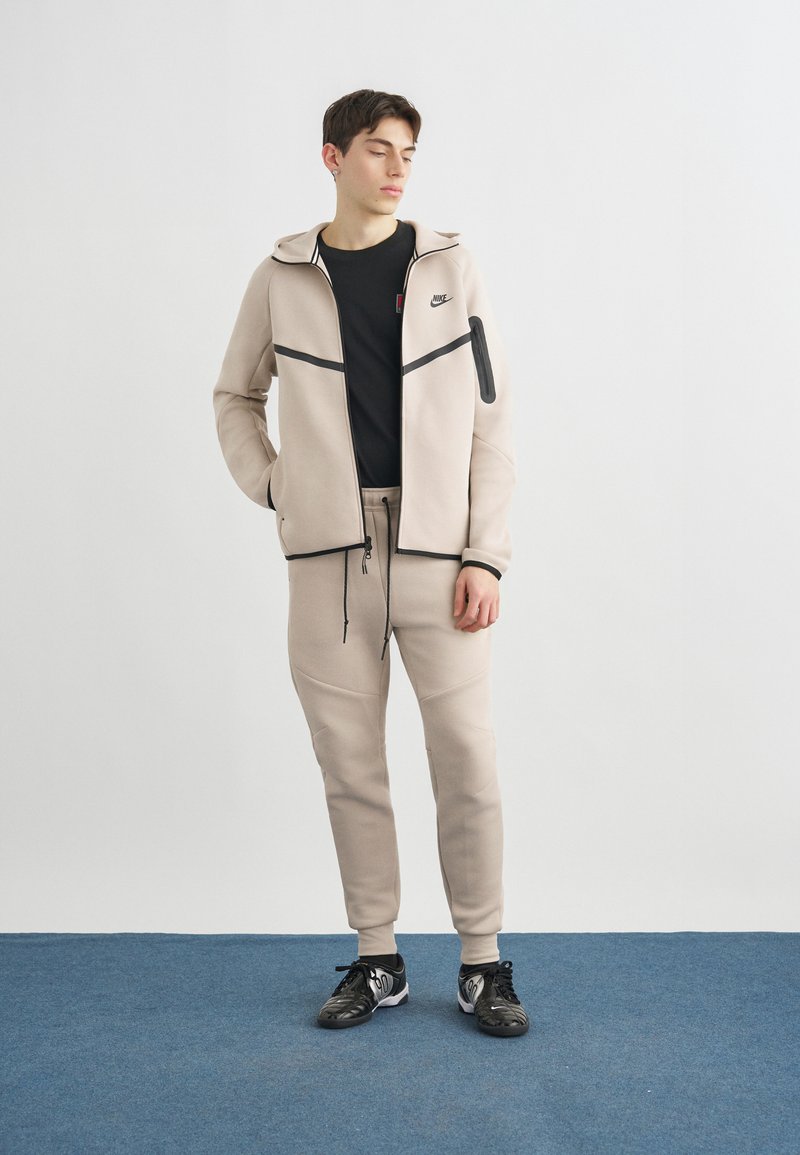 Joven de pie con los ojos cerrados, vistiendo un chándal beige de Nike, camiseta negra y zapatillas negras sobre un suelo azul frente a una pared blanca.