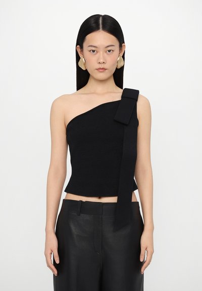 Top negro de un solo hombro con un tejido texturizado y un gran lazo como acento. Combinado con pantalones de piel negros de cintura alta.