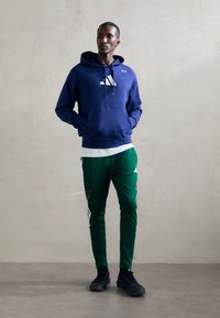 Námornícky modrá mikina s bielym logom, v kombinácii so zelenými teplákmi so bielymi a červenými akcentmi. Čierne športové topánky dotvárajú celý outfit.