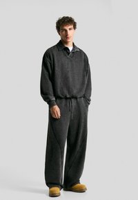 Giovane uomo in piedi con le mani in tasca, indossando una felpa grigia scura larga e pantaloni abbinati, e stivali beige, su uno sfondo chiaro e semplice.