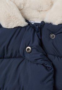 Doudoune bleu marine avec un col en fausse fourrure, dotée de boutons-pression argentés et d'une fermeture éclair. Texture lisse avec un détail matelassé.