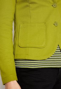Blaser texturé vert avec deux boutons et une poche plaquée, associé à une chemise présentant un motif à rayures noir et blanc en dessous.