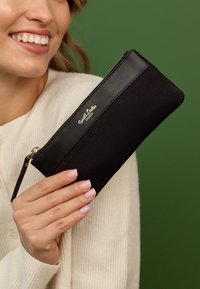 Mustast kangast tõmblukuga kott, millel on sile nahast aktsent ja kuldne logo. Omab randme rihma ja seda hoitakse käes rohelise tausta ees.