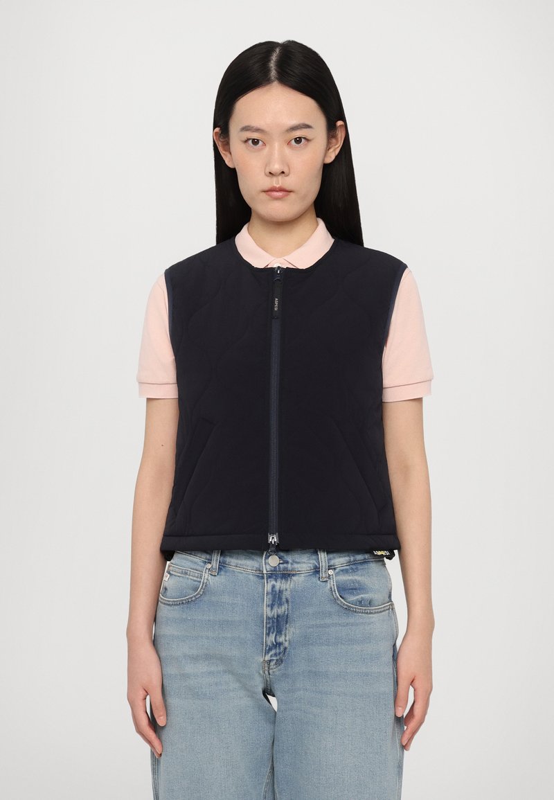 Quilted navy sleeveless vest with a front zipper. Styled over a light pink collared shirt, paired with light blue jeans.