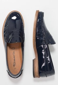 Mocassins en cuir verni bleu marine avec un talon en bois plat. Les caractéristiques incluent des coutures sur le dessus et une texture lisse et brillante.