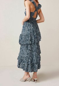 Robe maxi bleue à motifs floraux avec des bretelles à volants, un design à étages et une taille smockée. Associée à des talons ouverts de couleur claire.