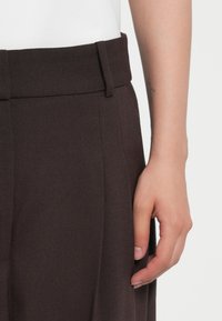 Pantaloni maro confeccionați, cu un talie lată. Țesătura este texturată, având plii și bucle pentru curea, prezentate alături de o mână.