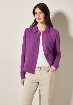 Camicia lunga viola con tessuto testurizzato, due tasche sul petto e chiusura con bottoni. Indossata sopra una maglietta bianca, abbinata a pantaloni beige.