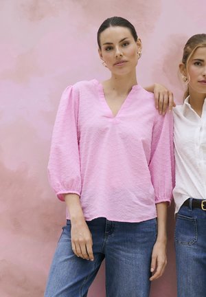 Dvi moterys stovi prie rožinės sienos; kairėje – ji dėvi rausvą tekstūruotą palaidinę ir mėlynas džinsines kelnės, dešinėje – baltą palaidinę ir mėlynas džinsines kelnes su diržu.