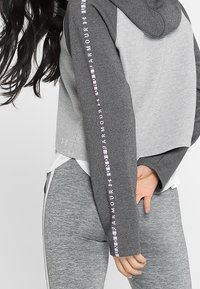 Haut à capuche court gris avec une texture rayée et une capuche. Doté d'un lettrage rose le long des manches. Associé à des leggings gris.