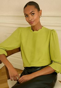 Limonkowa bluzka z falbanistym dekoltem, plisowanym przodem i bufiastymi rękawami, z szczegółowym wykończeniem na krawędziach rękawów.