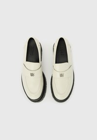 Mocassins en cuir blanc avec des semelles en caoutchouc noir, dotés d'une sangle sur le cou-de-pied et d'un logo en relief sur la semelle intérieure. Texture lisse.