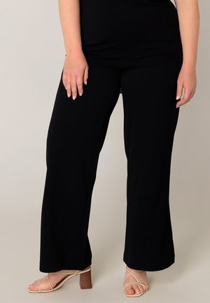 Persoon die zwarte wijde broek en beige sandalen met hak en bandjes draagt, staand tegen een effen lichte achtergrond.