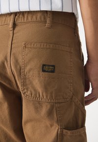Pantalones cargo marrones hechos de un tejido duradero, con un bolsillo trasero que lleva una etiqueta negra y detalles de costura visibles.
