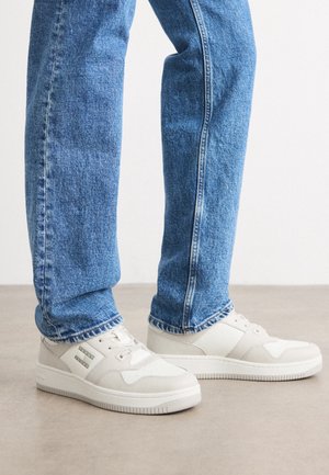 Beine in blauen Jeans und weißen niedrigen Sneakers, die auf einem glatten, hellen Boden vor einem schlichten Hintergrund stehen.