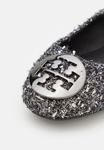 Svart och silver platt sko med en strukturerad, metallisk finish, med en rund, glansig logotyp emblem på tån.