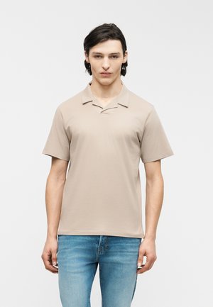 Junger Mann steht nach vorne blickend, trägt ein beiges Polo-Shirt mit kurzen Ärmeln und blaue Jeans vor einem einfarbigen hellen Hintergrund.