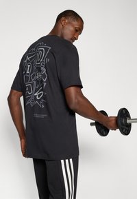 Schwarzes Baumwoll-T-Shirt mit grafischem Design auf der Rückseite, Rundhalsausschnitt, kurze Ärmel, kombiniert mit schwarzen Sporthosen mit weißen Streifen.