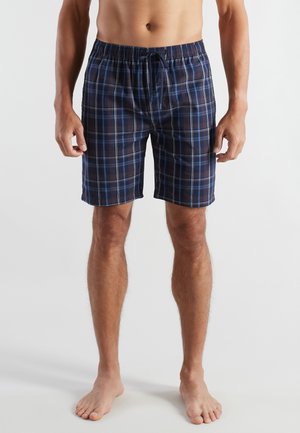 Mann trägt marineblaue, schwarze und hellblaue karierte Shorts mit elastischem Bund und Kordelzug, steht barfuß vor einem einfarbigen Hintergrund.