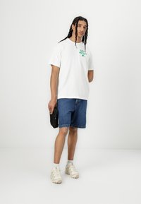 White cotton t-shirt featuring green graphic print, paired with blue denim shorts. Model holds a small black bag, wearing light-colored sneakers.