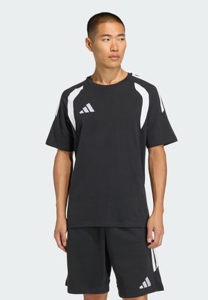Młody mężczyzna ubrany w czarną odzież sportową Adidas z białymi paskami, stojący z rozluźnionymi rękami, patrzący w prawo na jednolitym tle.
