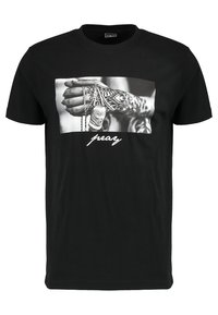 Black cotton t-shirt featuring a graphic print of hands with tattoos and chains. The word "pray" is displayed in white cursive text.