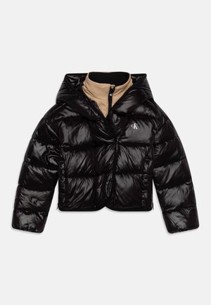 Giacca imbottita nera lucida con cappuccio, indossata sopra un colletto interno beige con zip, con logo bianco "cK" sul petto a sinistra.