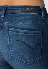 Vaqueros de mezclilla azul oscuro con un bolsillo trasero que presenta un sutil diseño de costura curva y una etiqueta negra marcada "ONLY". Textura suave, corte clásico.