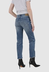 Nieprzydeptane niebieskie dżinsy z przetarciami na krawędzi, dwoma tylnymi kieszeniami i beżową etykietą na pasku, zestawione z czarnymi botkami na obcasie.