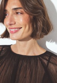 Top nero plissettato, con texture trasparente, scollo rotondo, maniche corte, indossato da una donna con carnagione chiara e capelli corti castani.