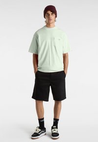 Mintgrünes Baumwoll-T-Shirt mit kurzen Ärmeln und einer Fronttasche, kombiniert mit schwarzen Shorts. Das Modell trägt eine kastanienbraune Strickmütze und schwarze Sneakers.