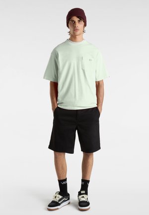 Camiseta de algodón verde menta con mangas cortas y un bolsillo en el frente, combinada con pantalones cortos negros. El modelo lleva un gorro de punto burdeos y zapatillas negras.