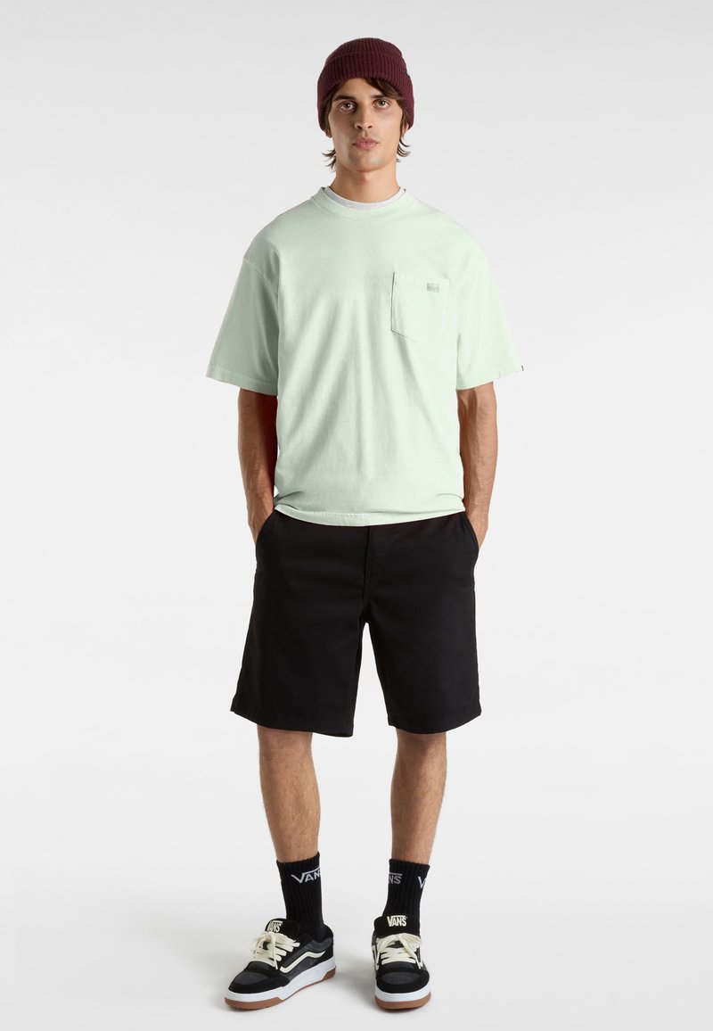 Mintgrön bomullströja med korta ärmar och en framficka, tillsammans med svarta shorts. Modellen bär en vinröd stickad mössa och svarta sneakers.