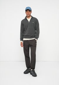 Junger Mann, der einen grauen Quarter-Zip-Pullover, schwarze Jogginghosen, dunkle Sneakers, ein weißes Poloshirt und eine hellblaue Baseballkappe trägt, steht vor einem weißen Hintergrund.