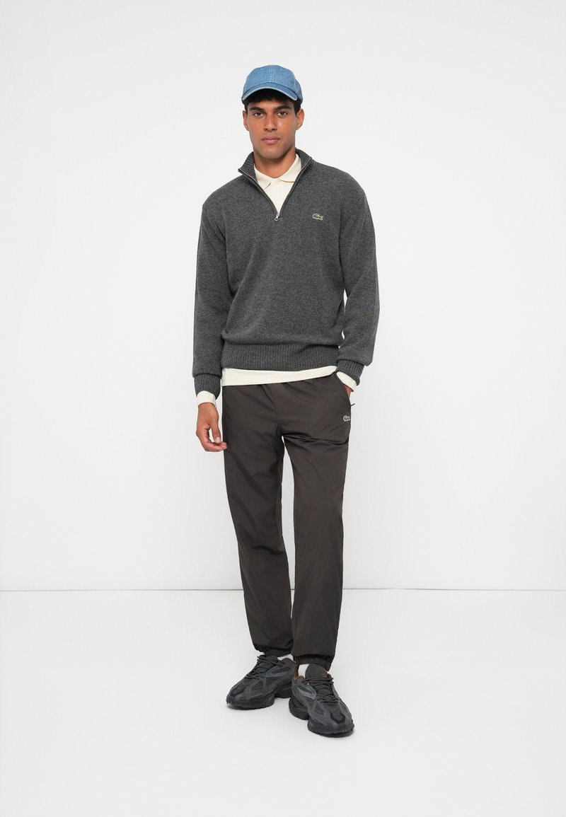 Ung man som bär en grå kvartslång tröja, svarta joggarbyxor, mörka sneakers, en vit polo skjorta och en ljusblå baseballkeps som står mot vit bakgrund.